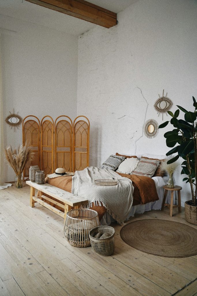 A bedroom with while walls and light wooden floor. A Bed with white, grey and orange covers. A gree plant. A wooden stand. 