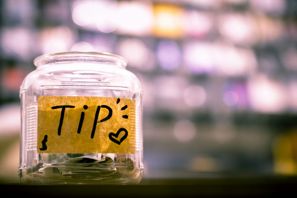 A tip jar with some coins in the bottom and yellow post it note saying TIP in black marker and a small heart, blurred background. 