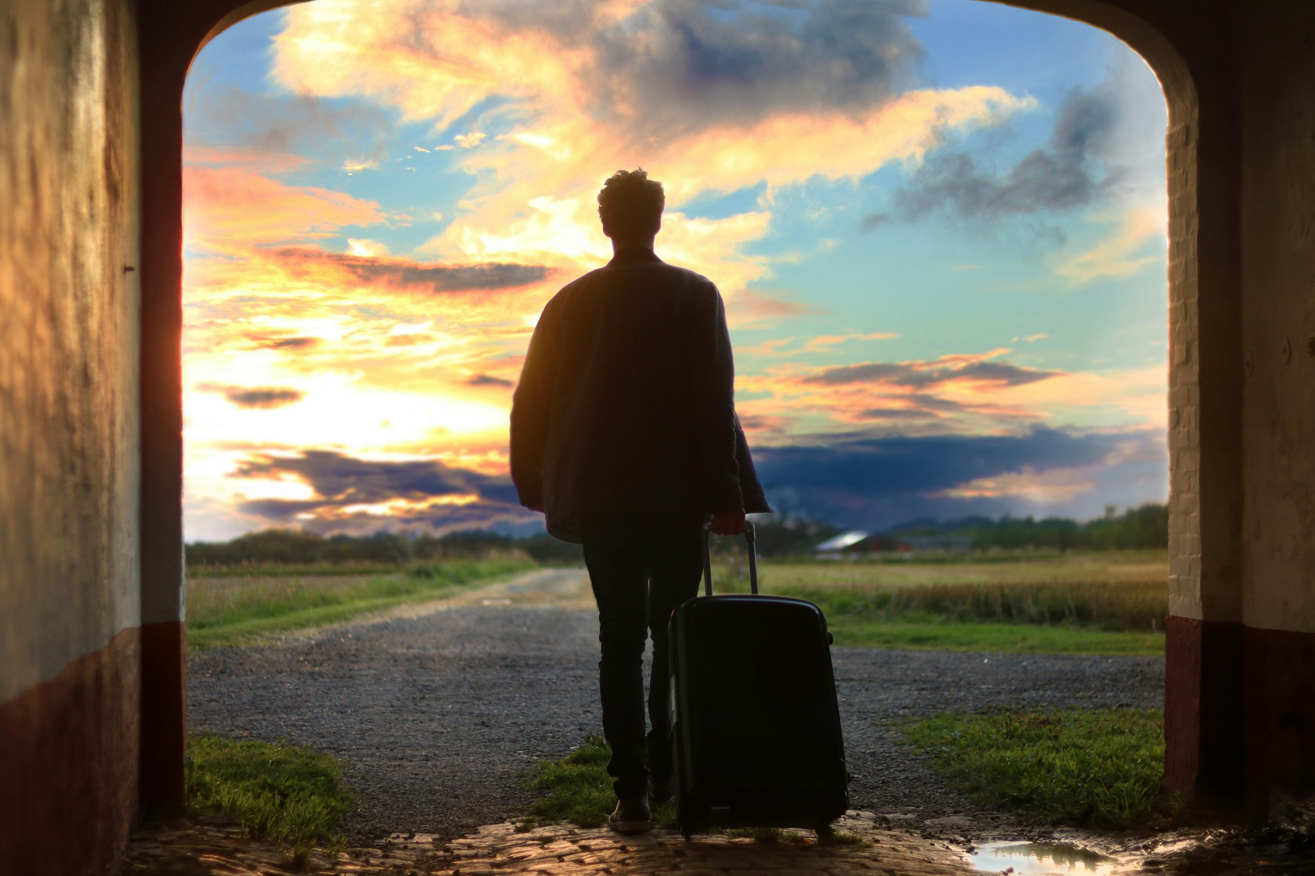 A man with a suitcase walking through a tunnel into a green area, blue sky with clouds, sunset 